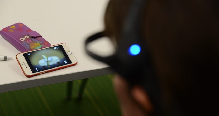 En mobiltelefon med en bild på en hjärna. I förgrunden en person med hörlurar på sig. Foto.