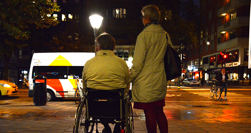 Person i rullstol med en assistent bredvid står vid en trafikerad gata.