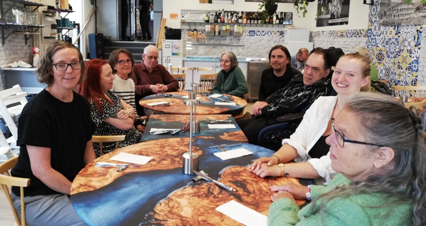 Ett tiotal personer sitter runt några sammandragna bord och tittar mot fotografen. Foto.