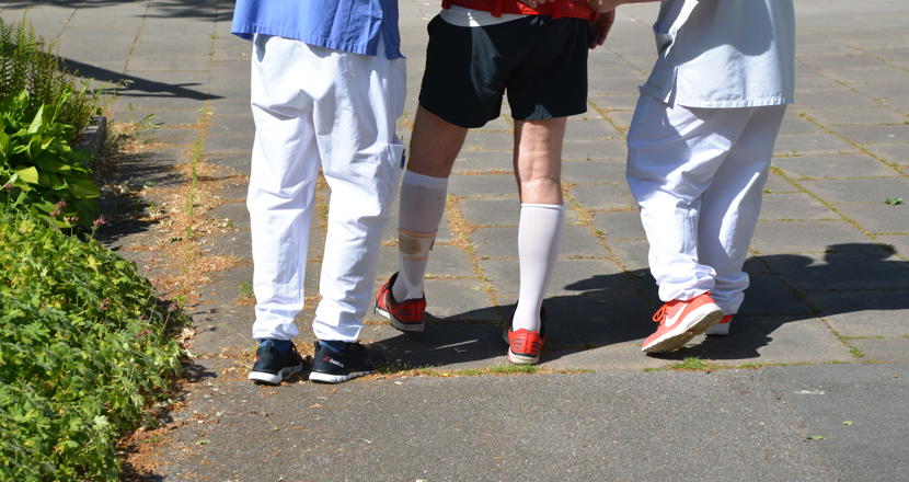 Två personer i vårdkläder som går på varsin sida om en person som gångtränar. Foto.