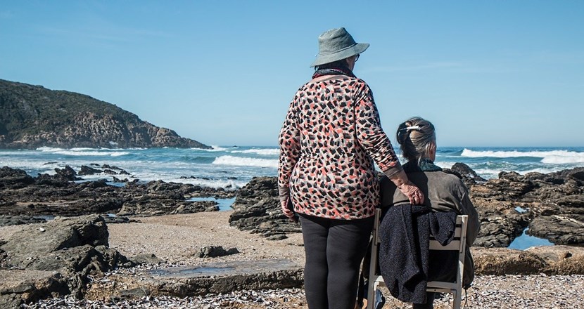 Två äldre kvinnor med ryggen mot kameran, på en stenig sandstrand. Foto.