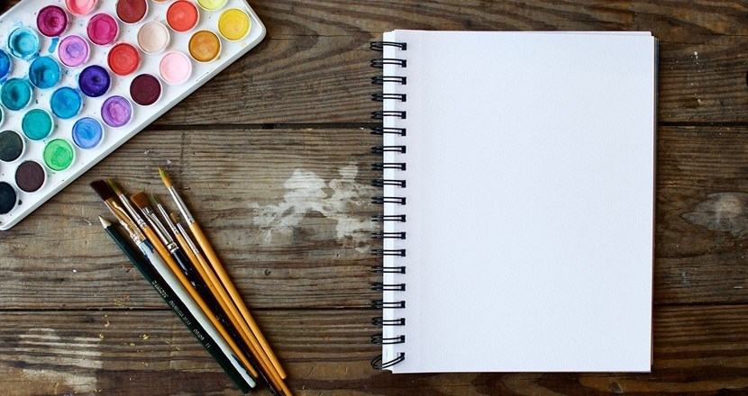 Akvarellfärger och penslar ligger på ett träbord bredvid ett block med en tom vit sida.