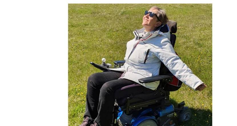 En lycklig Lise Lidbäck i sin elrullstol, i solen på en grön gräsmatta. Foto.