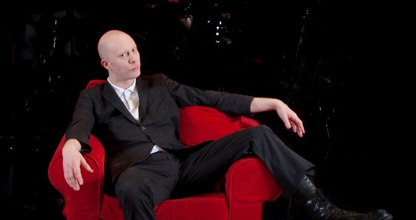 Programledare Loke Nyberg, som sitter slängd i en röd fåtölj, iklädd en svart kostym. Foto.