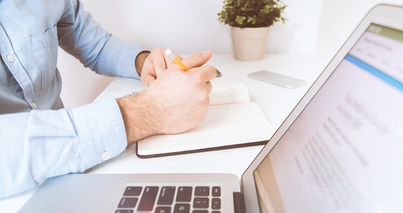 Fotografi på ett par händer tillhörande man som skriver i anteckningsblock, laptop i förgrunden. Foto.