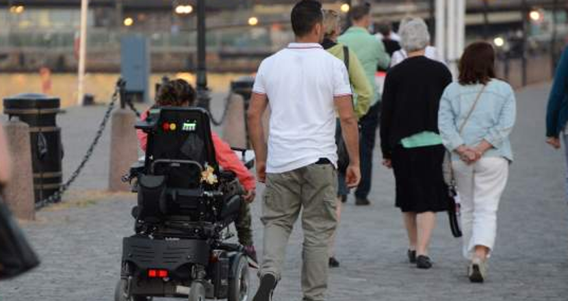 Person i elrullstol med man gående i intill, fotade bakifrån på promenad.