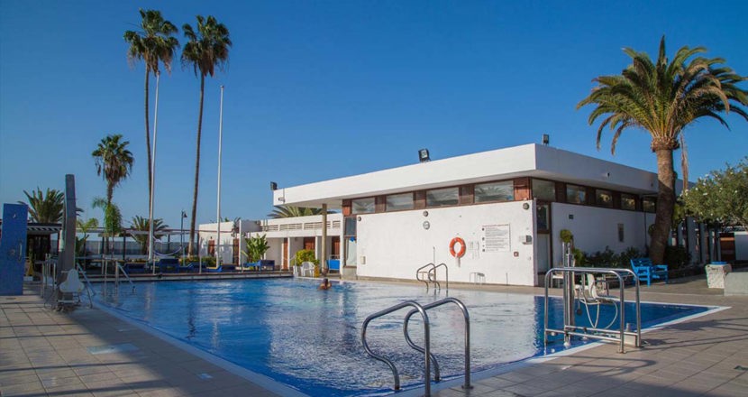 En vit byggnad mad en bassäng i förgrunden och i bakgrunden en blå himmel och några palmer. Foto.
