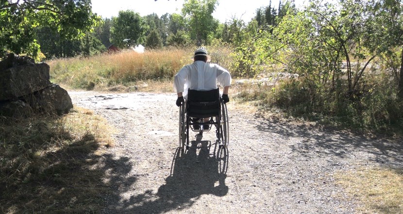 En person som kör rullstol på en liten, grusad väg genom gröna sommarhagar. Foto.