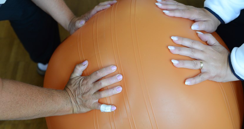 Närbild på händerna på en patient och en fysioterapeut som balanserar mot en orange pilatesboll. Foto.