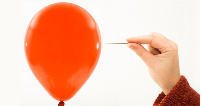 En person som sticker hål på en orange uppblåst ballong med en stor nål. Foto.
