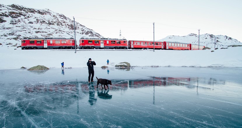 Ett långt rött tåg i ett landskap med snö och is. En man med en hund på isen i förgrunden.