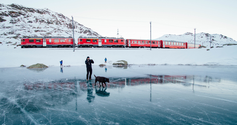 Ett långt rött tåg i ett landskap med snö och is. En man med en hund på isen i förgrunden.