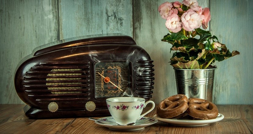 En gammal radio på ett vackert bord med en kaffekopp, ett fat med kringlor och en blomkruka. Foto.