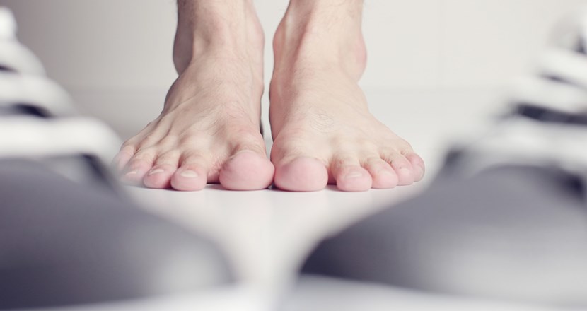 Två nakna fötter på ett golv med två suddiga skor i förgrunden. Foto.