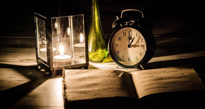 En ljuslykta med levande ljus, en väckarklocka och en uppslagen bok. Foto.
