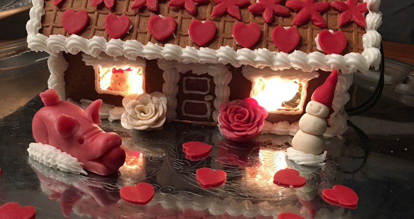 Pepparkakshus med invändig belysning. Framför huset finns en röd marsipangris, röda hjärtan, en röd och en vit ros samt en snögubbe i marsipan.