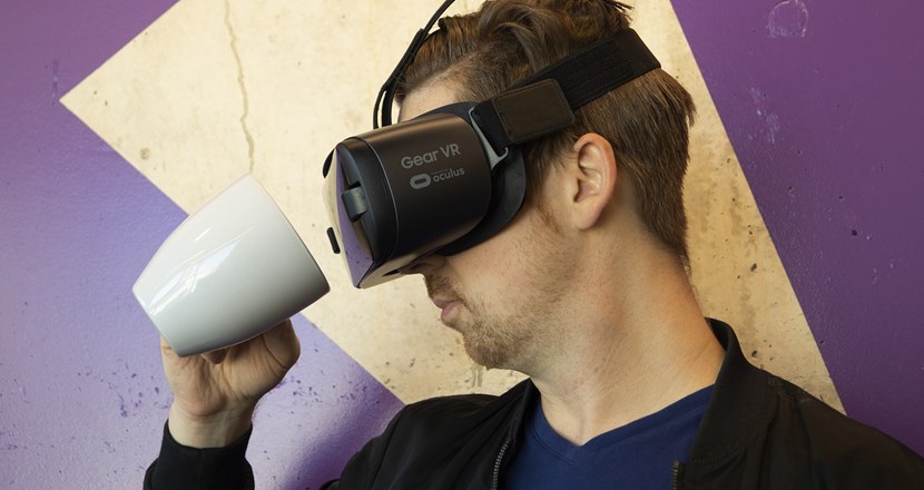 En man med VR-glasögon försöker dricka en kopp kaffe. Foto.