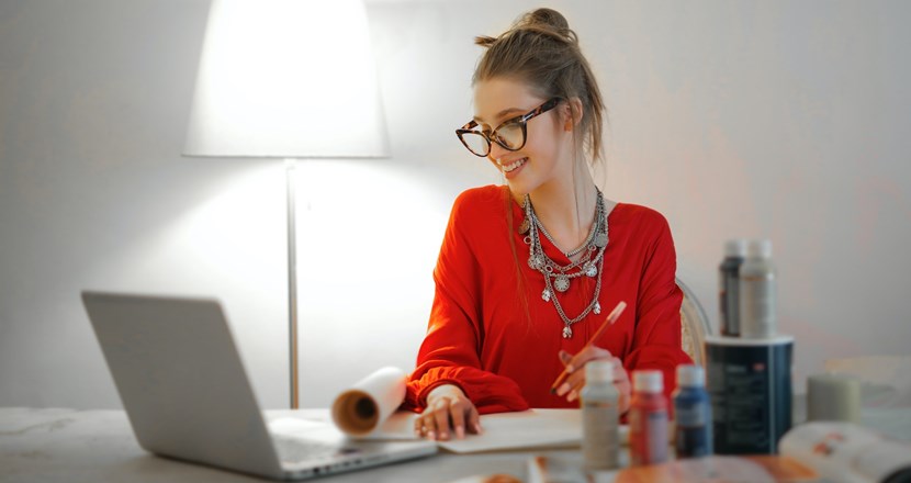 Ung kvinna i röd tröja som sitter vid ett bord med dator och deltar i ett digitalt möte. Foto.