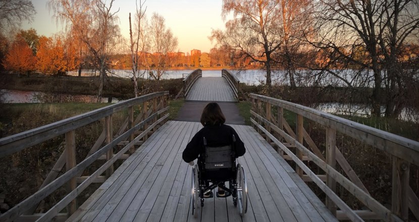 Emma bakifrån i sin rullstol på en träbro med vatten runt. Foto.