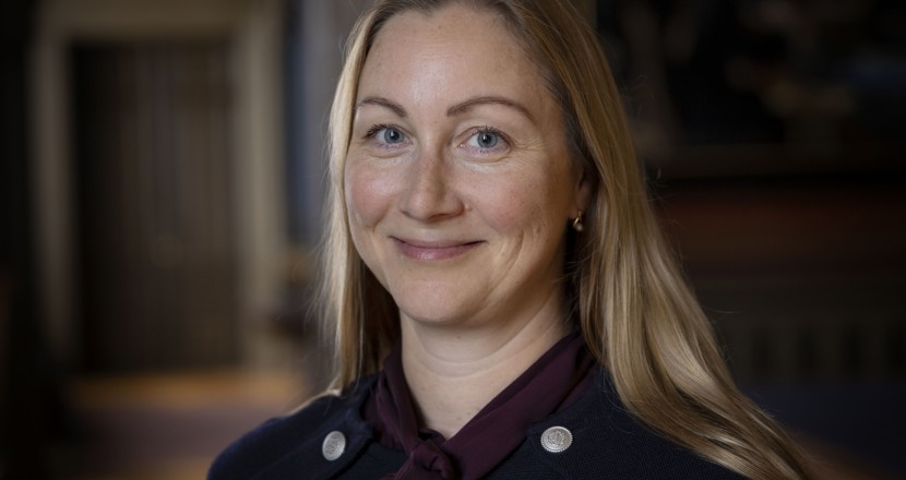 Anna har långt halvblont hår, blåa ögon och en mörk kavaj med blanka knappar. 