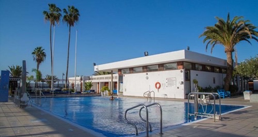 En av bassängerna och en av byggnaderna på rehabiliteringsanläggningen Vintersol mot en blå himmel och med några palmer i bakgrunden. Foto.