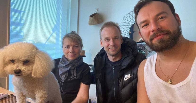 En bild med två män, en kvinna  i rullstol och en vit pudel sittandes vid ett bord. Foto.