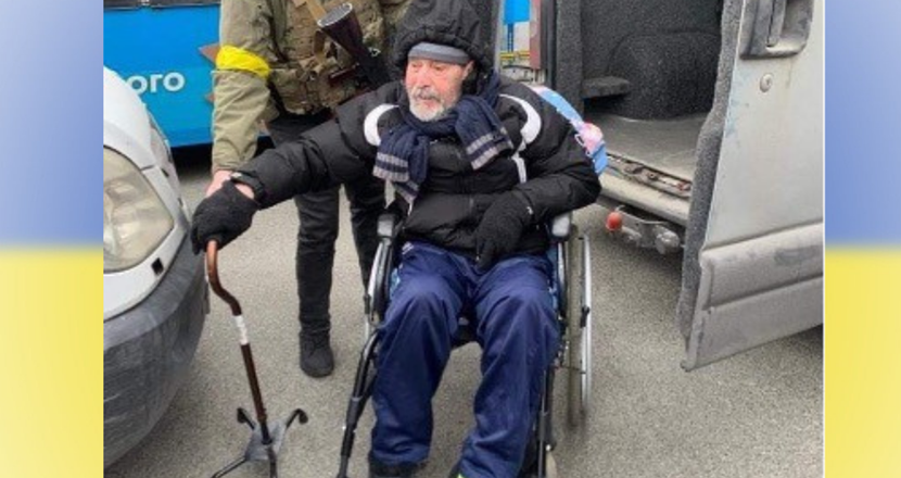 En man sittandes i rullstol hjälps av en soldat, med Ukrainas färger gult och blått som bakgrund. Foto.