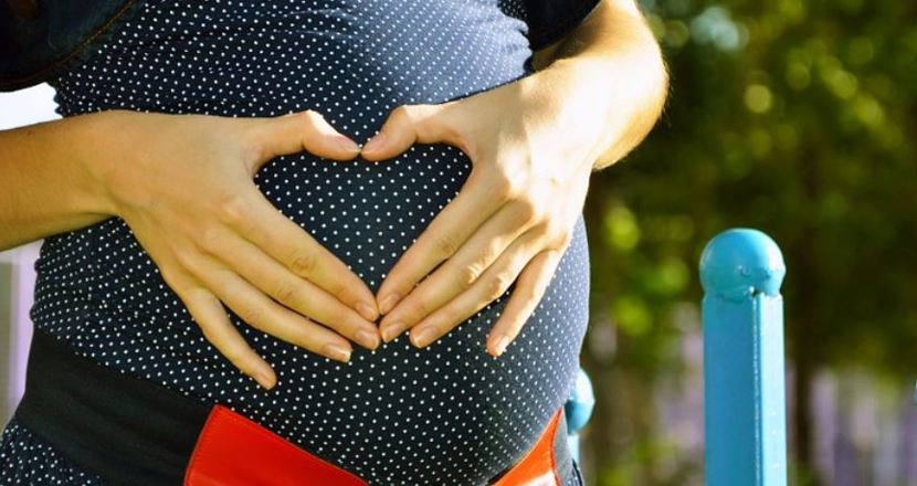 En gravidmage där kvinnan gör ett hjärta med händerna framför magen. Foto.