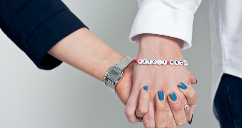 Närbild på två som håller varandra i händerna. En har ett guldarmband och ett armbandsur, den andra ett armband med texten 