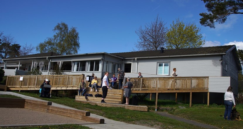 Flera människor på trappan till en träaltan framför ett grått lågt hus. Blå himmel och grön gräsmatta