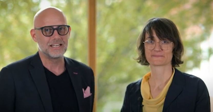 Henrik och Katharina har mörka kavajer och står framför ett stort fönster med grönskande träd i bakgrunden.