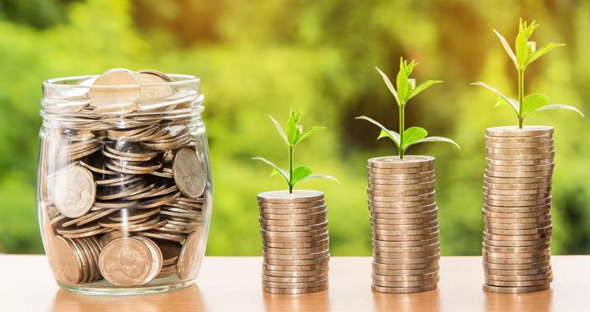 En glasburk och tre små travar med mynt framför en grönskande bakgrund. Foto.