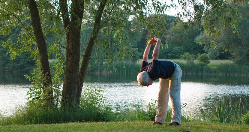 En äldre person som böjer sig framåt och stretchar, vid en sjö. Foto.