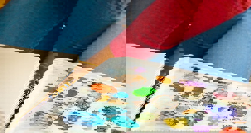 Färgprover av akvarell på ett vitt underlag samt knät på personen som målar i jeansklädda ben och röda tröjärmar. Foto.