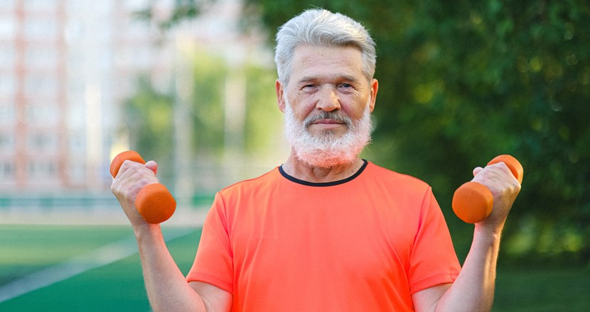 En man med grått hår och skägg och orange t-shirt som tränar med små hantlar utomhus. Foto.
