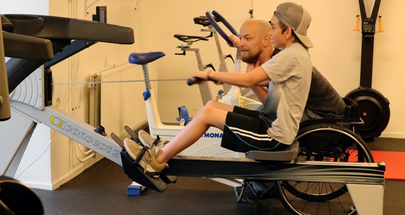 Tomas son tränar i en roddmaskin och Tomas peppar från sidan i sin rullstol. Foto.