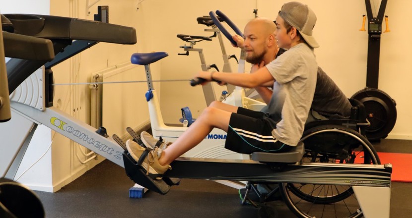 Tomas son tränar i en roddmaskin och Tomas peppar från sidan i sin rullstol. Foto.