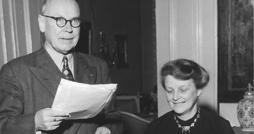 Svartvitt foto på en man och en kvinna. Mannen håller i ett papper. Kvinnan skriver på en skrivmaskin. Foto.