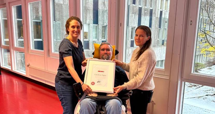 En man i elrullstol med ett diplom i knät. Flankerad av två kvinnor. Foto.