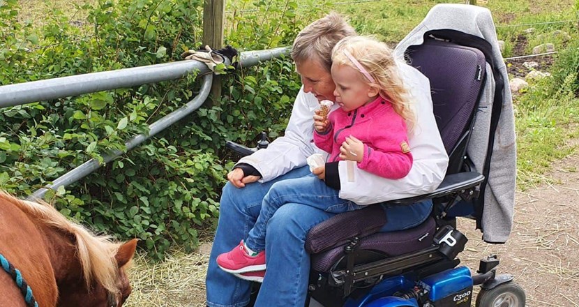 Lise i sin rullstol med ett barnbarn i knät. De äter glass och hälsar på en häst. Foto.