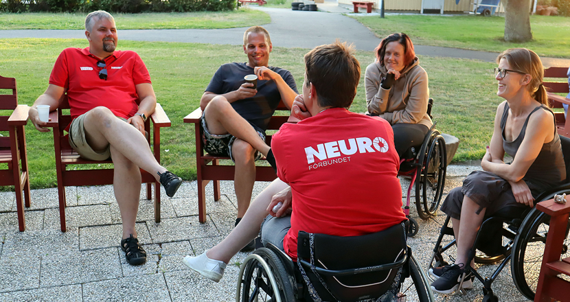 Fem person som sitter sommarklädda tillsammans och samtalar. Tre av dem i rullstol. Foto.