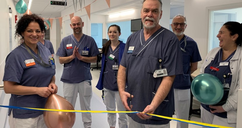 Sex personer i sjukvårdskläder framför ett blågult band. En man ska precis klippa bandet. Foto.