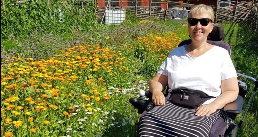 Sommarbild på Lise i sin rullstol med grönska och blommor i bakgrunden. Foto.