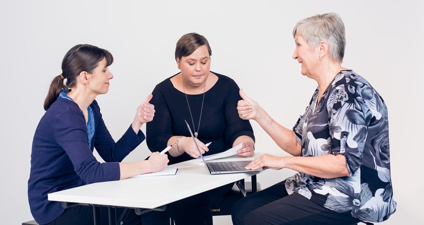 Tre kvinnor i olika åldrar pratar vid ett bord och gör tummen upp.