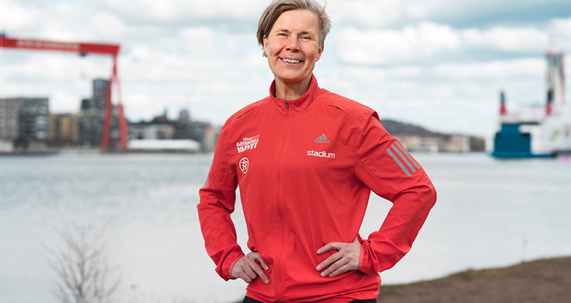 En leende kvinna i röd träningsoverallsjacka i förgrunden och i den suddiga bakgrunden vatten och en hamn. Foto.