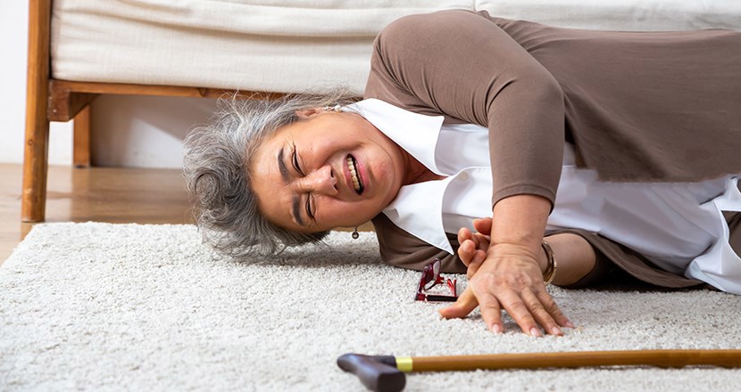 En kvinna som fallit ligger på golvet och sträcker sig efter sin käpp. Foto.
