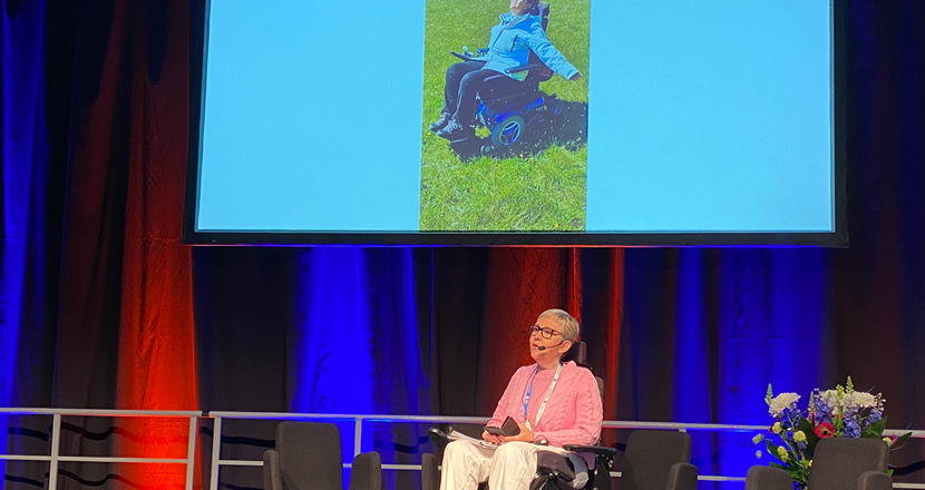 Lise Lidbäck på scenen. I bakgrunden en bild på en lycklig Lise i sin el-rullstol - på en äng med gullvivor. Foto.