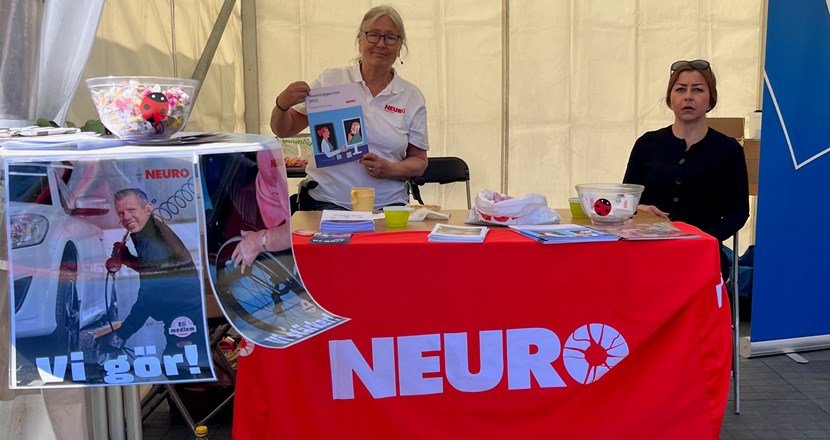 Ett bord med röd duk och Neuros logotype i vitt. På bordet ligger broschyrer och bakom bordet står en person och en som sitter.