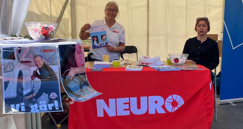Ett bord med röd duk och Neuros logotype i vitt. På bordet ligger broschyrer och bakom bordet står en person och en som sitter.
