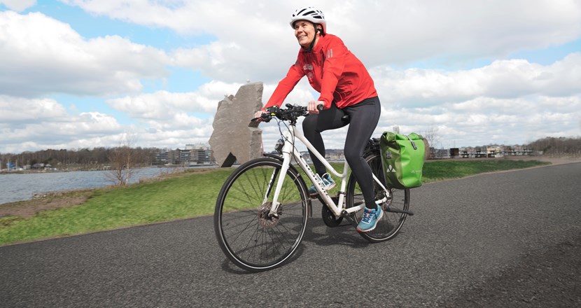 Jenny i vit cykelhjälm cyklar på asfaltväg, på vänster finns en sjö